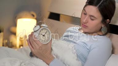 女孩设置闹钟。 一个年轻漂亮的女人独自躺在卧室里躺在床上，手里拿着一个闹钟